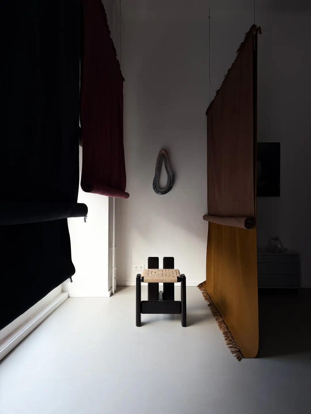 Rugs in black, burgundy and ochre hang from the ceiling of the white studio, framing a woven stool