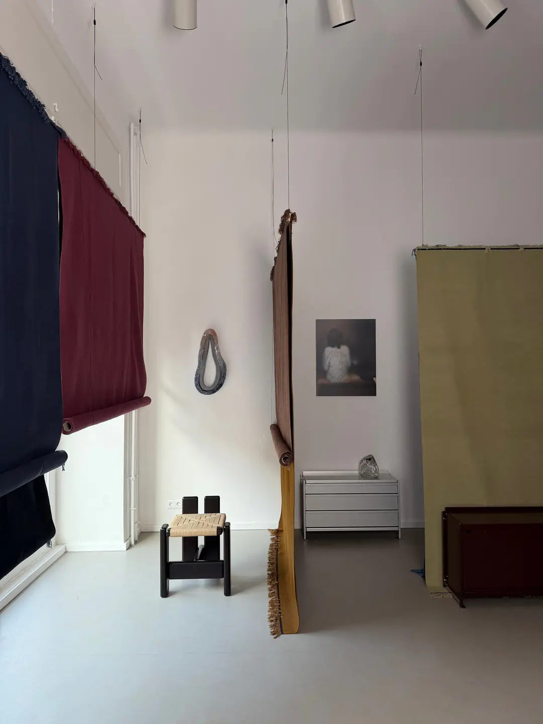 The installation: rugs in navy, burgundy, ochre and olive hung from the ceiling, with a woven stool, an aluminium chest and a photograph