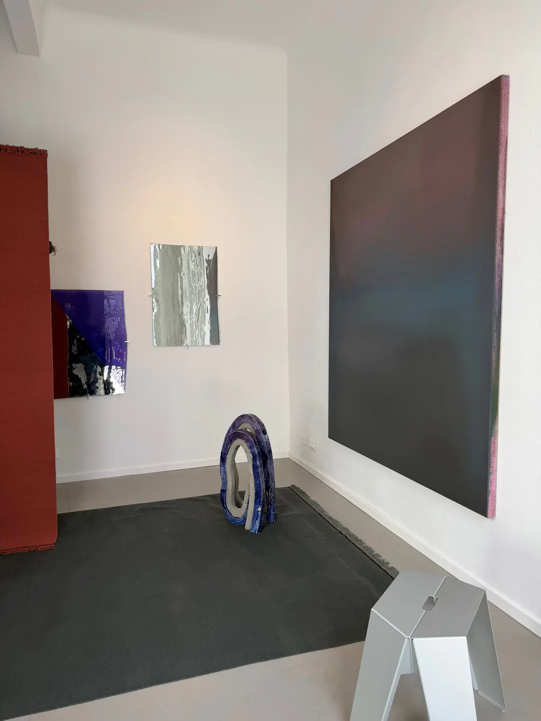 A large dark canvas, a blue ceramic sculpture and silver works on paper around a grey rug